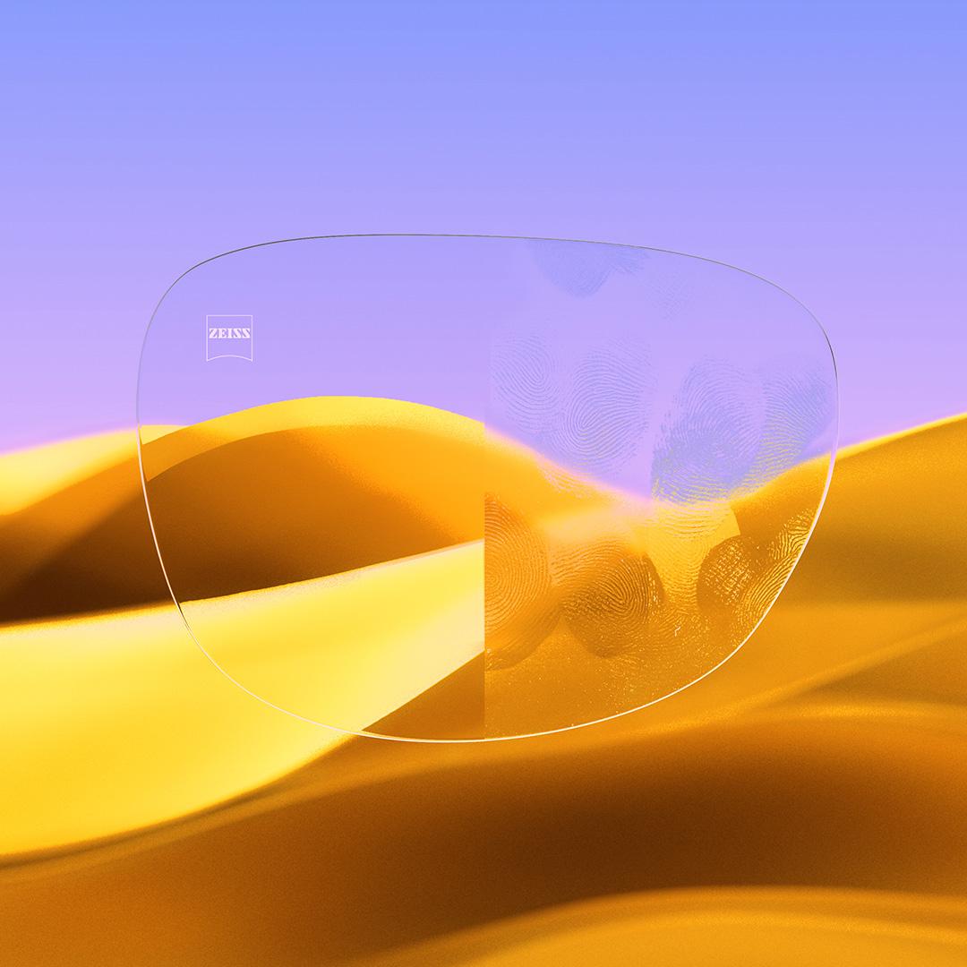 Egy szemüveglencséről készült osztott nézet: az egyik fele átlátszó és tiszta, ZEISS DuraVision Gold UV réteggel ellátva, míg a másik fele tele van ujjlenyomatokkal.