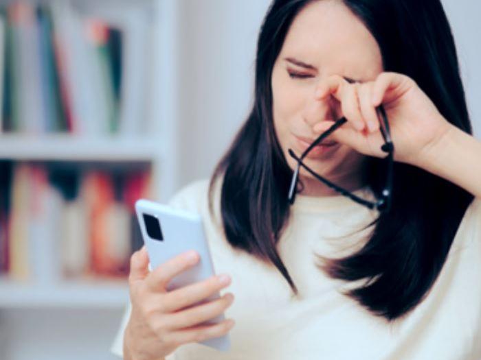 Egy kép, amelyen egy 30-as éveiben járó nő a telefonját nézi, miközben a szemét dörzsöli.