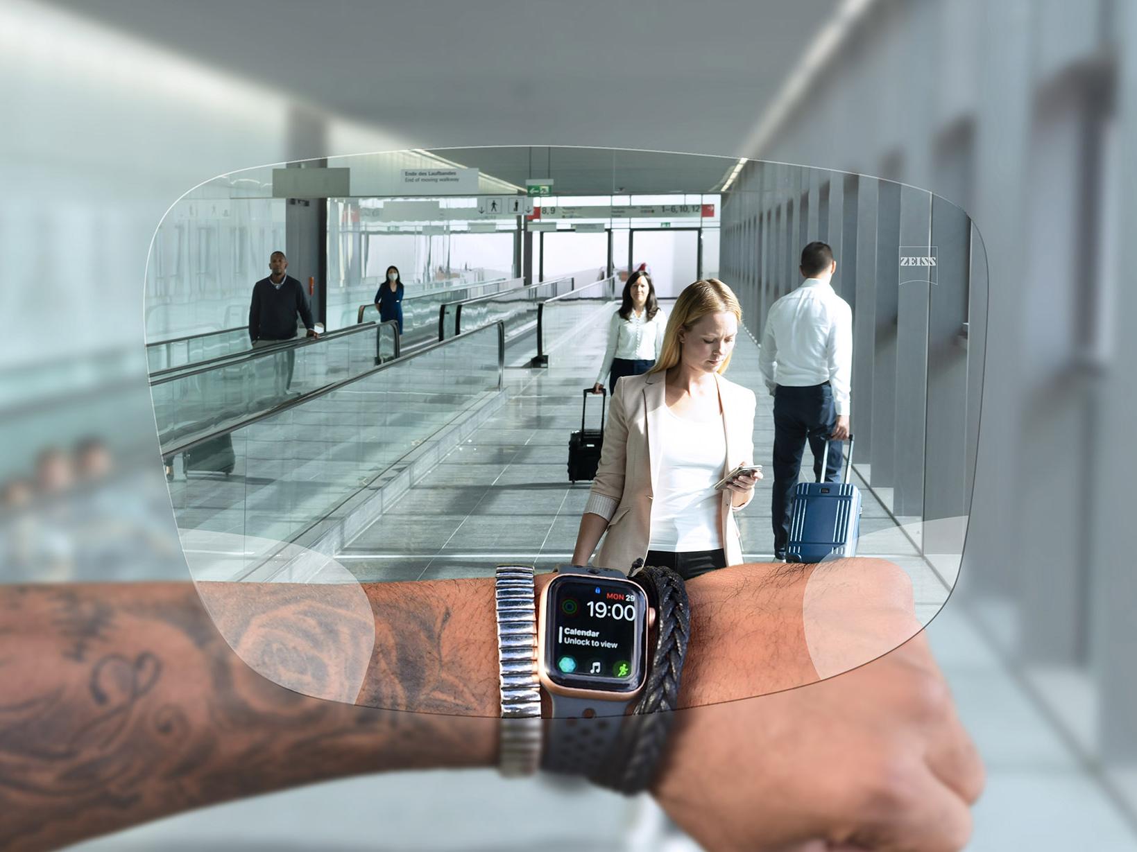 Az órájára ZEISS SmartLife Digital lencséken keresztül pillantó személy. A háttérben egy tipikus repülőtéri szituáció látható, emberek sétálnak a mozgólépcsőkön, és tolják a bőröndjeiket. 