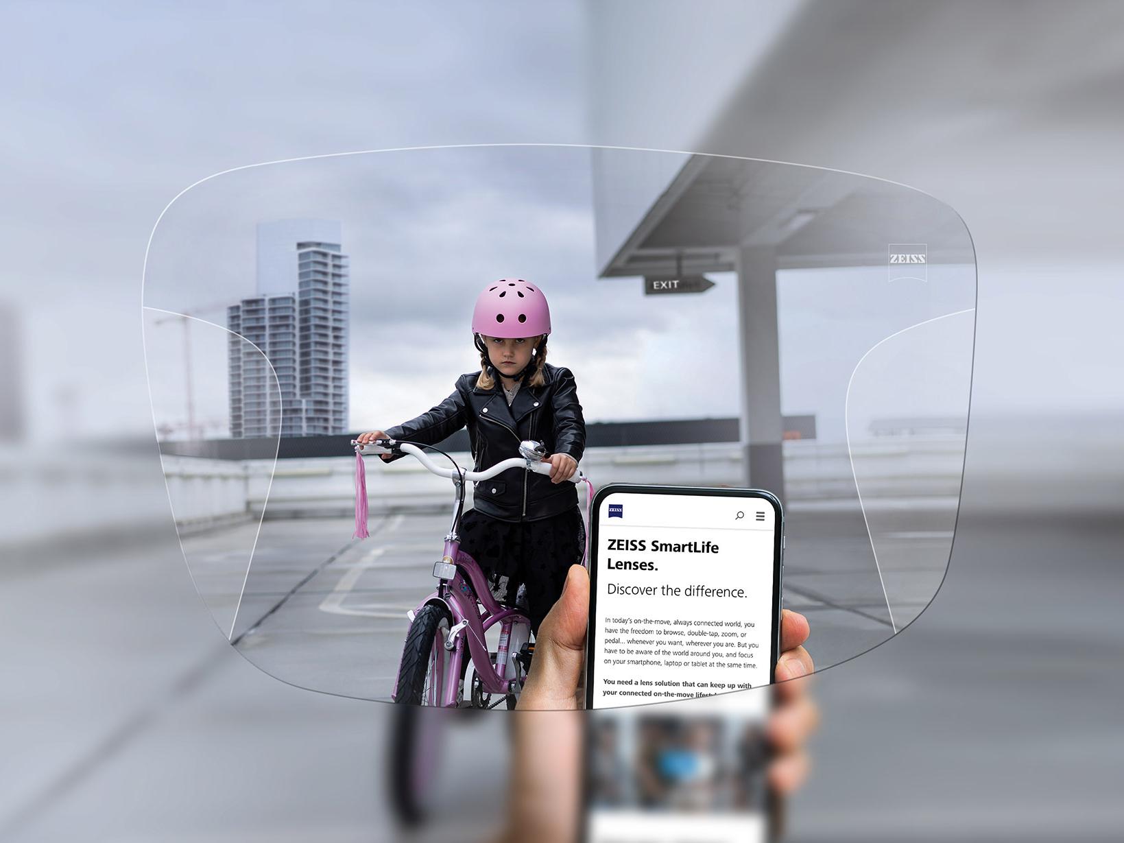 A telefonját a ZEISS SmartLife Progressive lencsén keresztül néző személy. A háttérben egy kerékpározó lány látható, aki rózsaszín bukósisakot visel. 