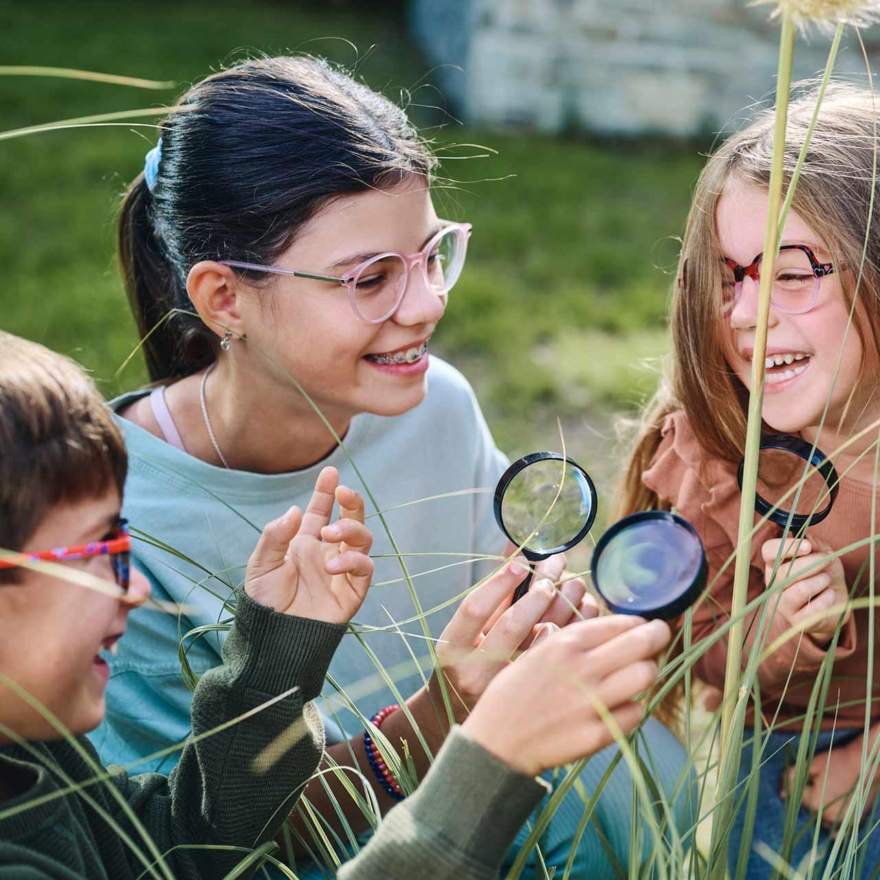 Szemüveget viselő gyermekek csoportja, akik nagyítókkal játszanak a szabadban és nevetgélnek