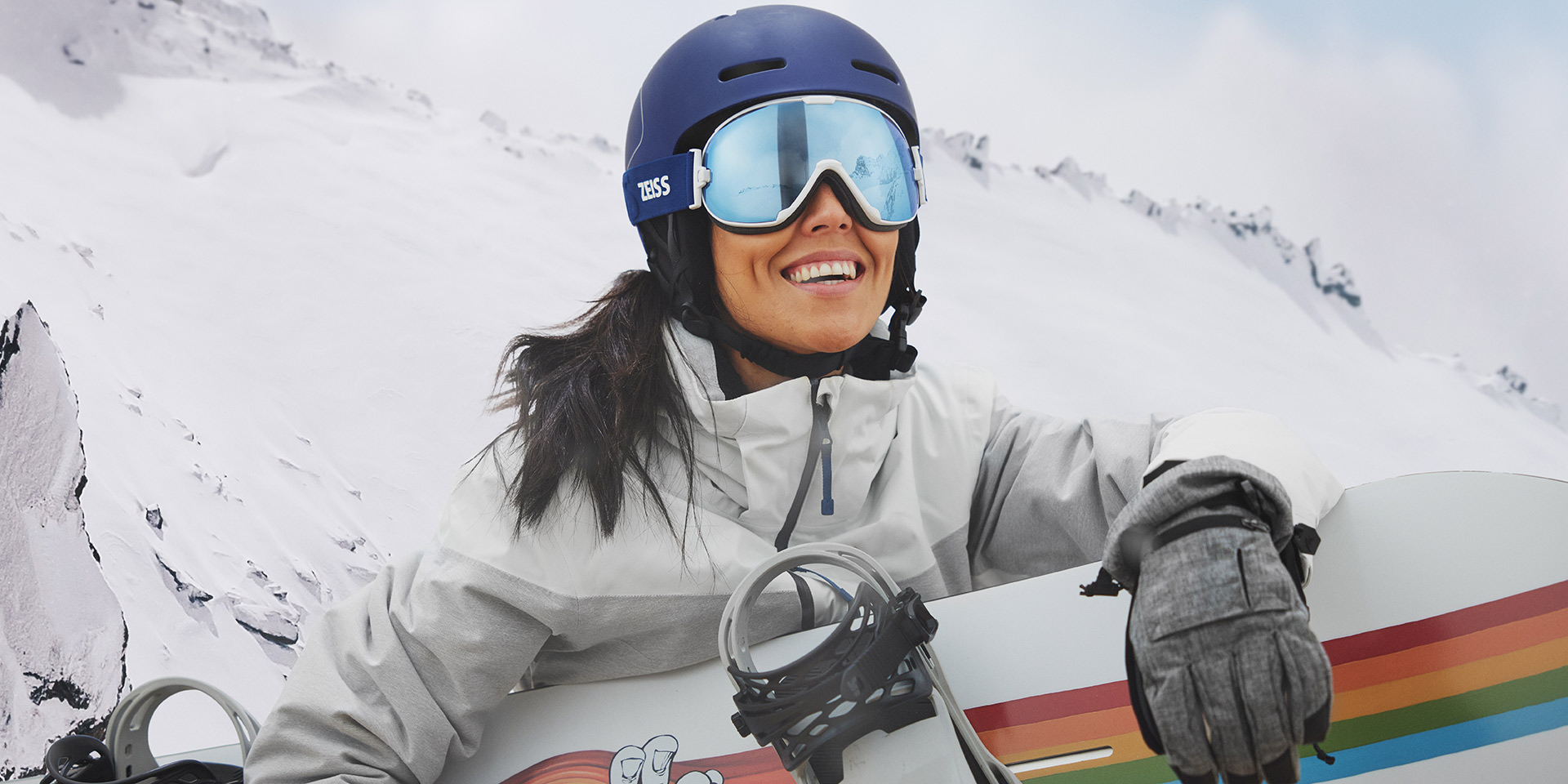 Snowboardot tartó és ZEISS cserélhető lencsés technológiával ellátott síszemüveget viselő fiatal nő.