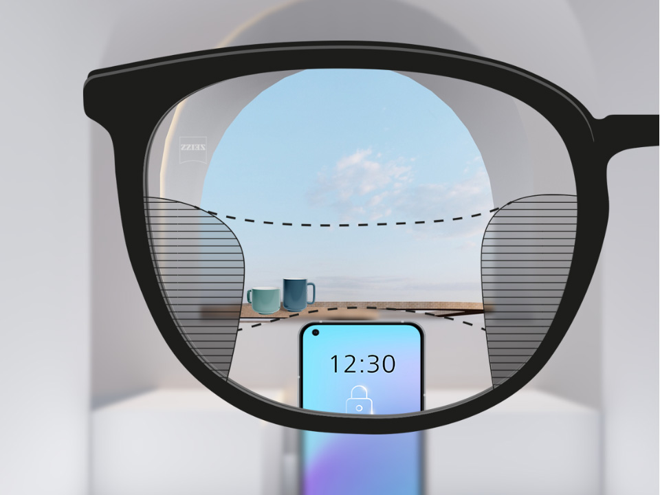 Saját szemszögből készült kép egy ZEISS SmartLife multifokális szemüveglencséről, amelyen keresztül élesen látszik a háttérben az okostelefon és két bögre is; a lencse bal és jobb oldalán pedig apró homályos részek látszanak.