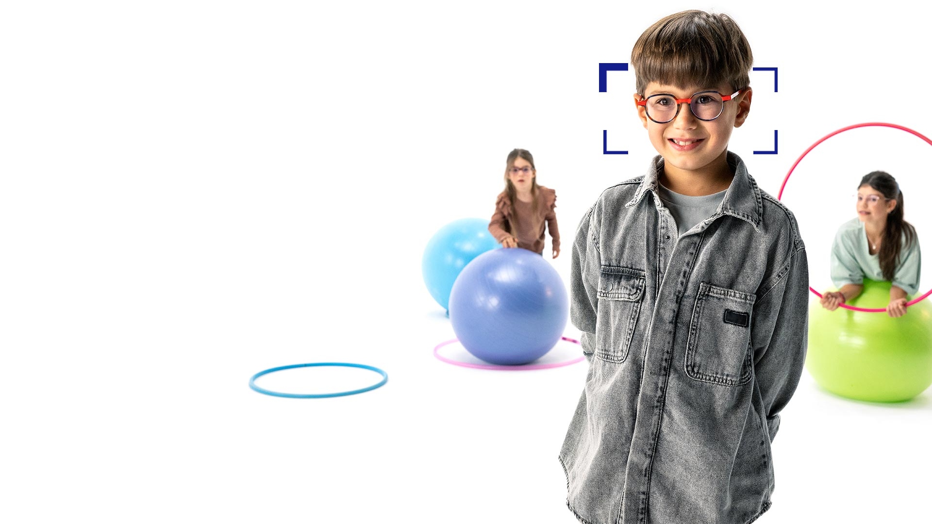 Barna hajú, kerek szemüveget és ZEISS MyoCare szemüveglencsét viselő kisfiú, aki az előtérben áll, és a kamerába mosolyog. A háttérben két ZEISS MyoCare szemüveglencsét viselő kislány játszik hulahoppkarikákkal és gimnasztikai labdákkal.